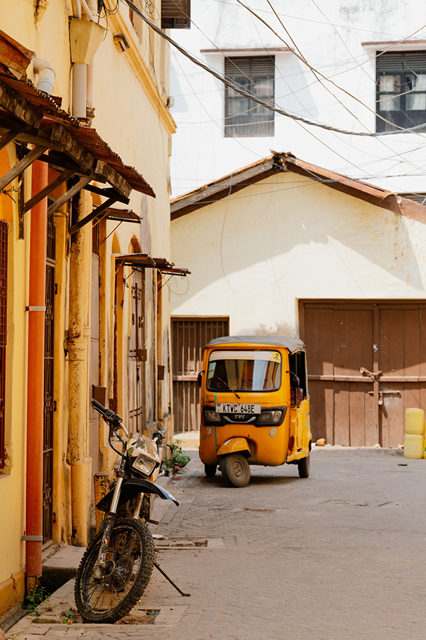 A weathered yellow building with wooden shutters lines a narrow cobblestone street in Mombasa. An orange and black auto-rickshaw is parked in the center of the quiet street, while a motorcycle leans against the yellow wall on the left. Wooden gates and additional colonial-era architecture are visible in the background. The scene conveys a sense of nostalgic charm and local authenticity.